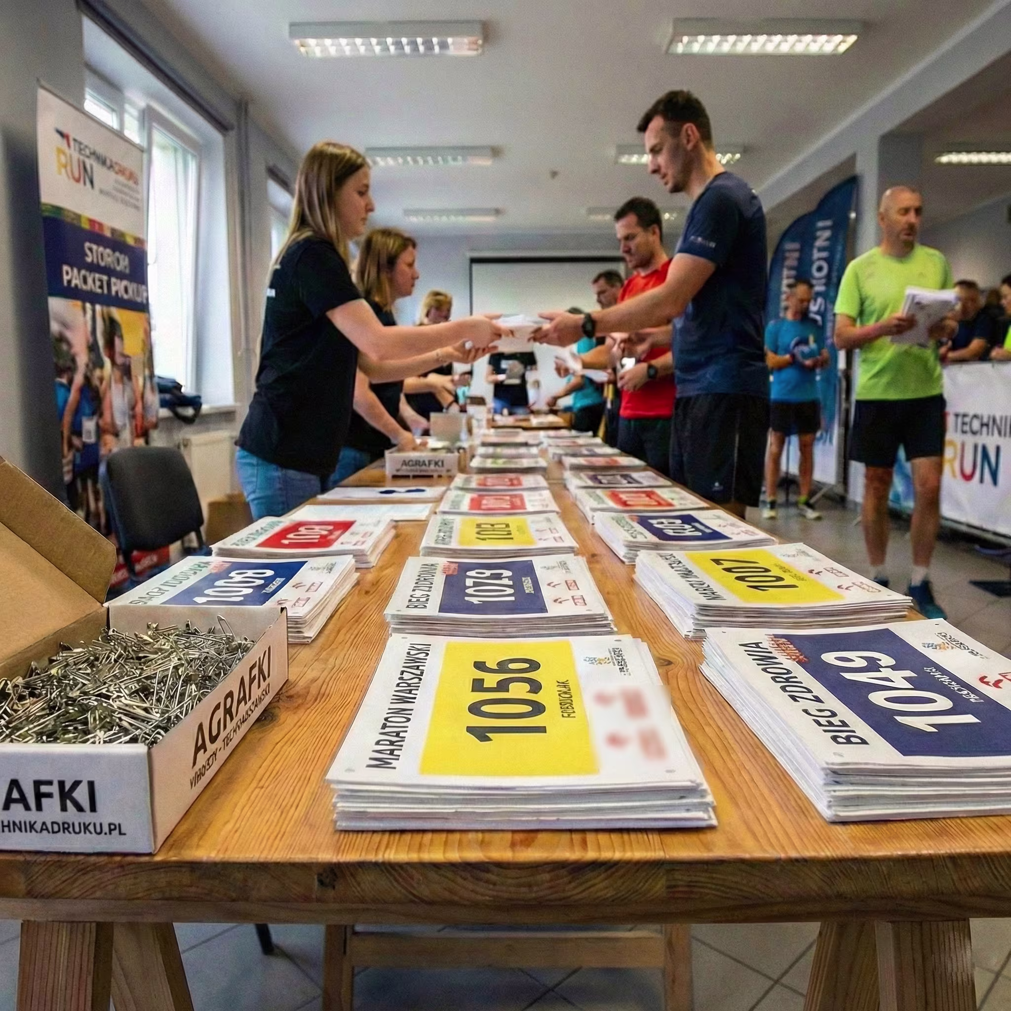 Biuro zawodow Biuro zawodów sportowych: na drewnianym stole leżą uporządkowane stosy personalizowanych numerów startowych w kolorach białym, żółtym i niebieskim. Na pierwszym planie otwarty karton z agrafkami oznaczony napisem 'AGRAFKI 1000 SZT'. W tle obsługa wydaje pakiety startowe zawodnikom.